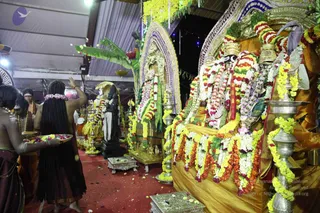 A photograph of a temple-style ritual hall with a red floor scattered with flower petals. On the left, a person with long dark matted hair, bare-backed and wearing a maroon wrap, walks away carrying a brass plate of flowers, while another attendant in saffron-toned clothing stands behind. At right, a large ornate golden throne-like seat is heavily decorated with white, yellow, orange and red garlands, and a golden canopy frames a deity murti. Brass lamps with garlands stand nearby, and a smaller dark statue sits at center beside a green banana-leaf backdrop. Petals fall through the air.