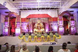 A photograph of a decorated stage in a hall with purple-lit pillared arches, a red tiled canopy and a white curtain backdrop. At the centre sits a large gold-decorated seated deity murti adorned with flowers, flanked by smaller figures. Yellow marigold garlands drape the stage edge, with white flower arrangements and green foliage below. Several seated people in the foreground, including a woman in white with a dark rudraksha-style necklace and a woman in orange, watch and photograph the stage with mobile phones. No speaker is clearly visible on the stage.