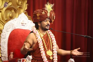 A man in a rust-brown turban with a yellow-green feather ornament and gold brooch, wearing a rust-brown robe, a long gold chain with a large pendant, pink-and-white flower garlands, and rudraksha bracelets, gestures with both open hands while speaking into a microphone. He sits in front of an ornate white-and-red throne with carved gold panels behind it and a red curtain. Swamiji (probable): the turban, ornate gold jewellery, garlands and rudraksha cues fit the page's Swamiji attribution, so he is listed as an in-person candidate. A copper vessel and small objects sit on the table at lower left.