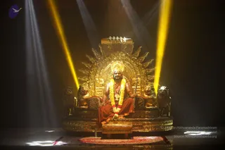 A seated man in saffron-orange robes sits on an ornate golden throne with lion-head armrests and a radiant sunburst backrest, lit by bright yellow spotlight beams in a darkened hall. He wears a red-and-white flower garland, a crown-like headpiece, and a rudraksha-style necklace, holding a staff in his right hand. A red patterned rug lies at the base of the throne. Swamiji (probable) appears as the seated figure, based on the saffron robes, ornate crown and flower garland cues. Text in the lower right reads the copyright line and archive credit.