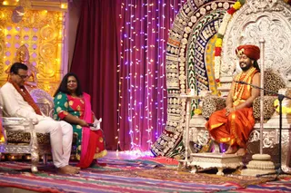 A satsang stage with a man in white kurta and a woman in green and pink sari seated on silver chairs at left, the woman holding a microphone. At right, a bearded man with a red-orange turban, orange silk dhoti and shawl, rudraksha-style garland and gold necklaces sits on an ornate silver throne with leopard-print cushions, a microphone stand before him, smiling. Behind is a decorated backdrop of draped fabric, fairy lights, a golden shrine with a deity murti at left, and marigold garlands on the throne. Colourful striped rug covers the floor.