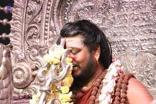 A photograph of a bearded man with long dark hair, eyes closed, bowing his forehead against a silver trident (trishul) decorated with a garland of yellow marigolds and pink flowers. He wears a rudraksha mala and a ceremonial gold necklace over a reddish-orange garment, with white and red markings on his forehead. Behind him is an ornate embossed silver backdrop with repeated figures and floral scrollwork.