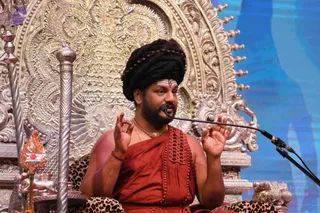 A man seated on an ornate silver throne with carved floral and scrollwork backrest, speaking into a headset microphone with both hands raised in mudra gestures. He wears long swept-back dark hair with a rudraksha band, a red-orange draped garment with decorative pleating, a gold chain necklace, and a red bead bracelet. He has a tilak mark on his forehead. Beside him stands a silver staff and a small silver ritual vessel with red decoration. Behind him is a blue backdrop. Probable Swamiji (long hair, rudraksha, saffron clothing and gold necklace) seated as the speaker at a satsang.