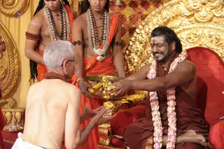 A seated man in a dark maroon robe with a pink-and-white flower garland, a rudraksha mala, and a vertical tilak on his forehead sits on a red cushioned throne with ornate gold carved panels, smiling as he reaches out to take bananas from a large brass bowl. Two young attendants in orange dhotis and white beaded necklaces stand behind, one holding the bowl. In the foreground an older man with grey hair, glasses and a bare back kneels with his palms extended toward the offering, wearing a white sacred thread and a rudraksha mala. The setting appears to be a ritual offering or darshan with banana fruit offerings.