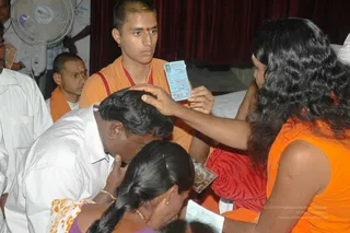 A darshan blessing scene in a hall with a red curtain and a standing fan at upper left. A long-haired figure in an orange sleeveless top and dark loose hair holds a printed card near the head of a man in a white shirt who bows forward with his head lowered. A young monastic in saffron robes with a red shoulder cloth and a red bindi stands behind, watching. A woman in a purple top with a braid bends forward in front, and another man in white sits at left with an orange shoulder cloth. The card is a printed blessing slip, partially legible.