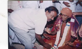 A photograph of a seated figure in orange robes and an orange turban with a red tilak, wearing a white jasmine garland with blue and red accents, a long rudraksha mala and a gold pendant, and a purple-and-gold shawl. A man in white shirt and white dhoti bends forward and touches the seated figure's hand with his own, while other people in white stand behind. Scattered flower petals lie on the floor.