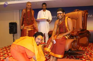 A photograph of a seated man in a brown leather armchair with carved wooden armrests, wearing a maroon and gold-bordered robe, a rudraksha mala with multiple beads, and a white tilak on his forehead, smiling while holding a small white card. A younger man in saffron-orange robes kneels at his feet on a red and white patterned carpet, also with a tilak. Behind them, a man in orange-maroon robes with a long rudraksha mala stands beside a man in white kurta holding a tray with a red cloth. A black speaker stands on the left, and a blue backdrop with partial lettering is behind them.