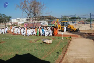 Outdoor photograph of a procession across a landscaped lawn at a construction-stage ground. A line of roughly 40-60 people, mostly in white clothing with some in saffron/orange robes and a few in green, blue and pink saris, walks along a path past a small rectangular pond with a large boulder. A yellow JCB backhoe loader stands at right beside a decorated colourful canopy or float, and a construction frame of blue steel scaffolding stands behind. Bare trees, a multi-storey white building and clear blue sky are visible in the background. No identifiable a person figure is clearly visible in this view.