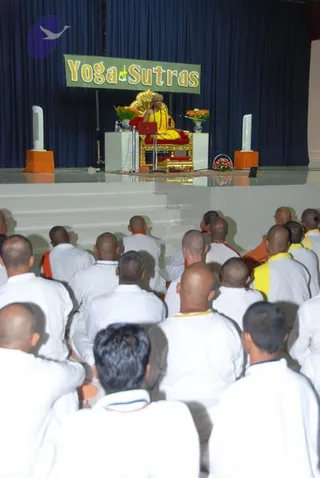 Photo of a Yoga Sutras satsang hall with a large green banner reading "Yoga Sutras" above a stage. A figure in yellow robes sits on an ornate gold-and-red throne behind a small laptop and a flower arrangement with orange blooms. Two white speaker columns flank the stage. In the foreground, a large seated audience of shaved-headed men in white clothes with yellow and orange sashes faces the stage, with their backs to the camera. Roughly two dozen people are visible.