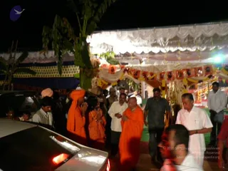 Night photo of an outdoor festival area at a decorated pandal with white draped canopy, yellow and orange marigold garlands and banana plant. A bald man in an orange saffron robe with hands folded in namaste walks among a crowd; a figure in orange with an orange turban and another in orange stand nearby, and men in white shirts and dhotis and a man in grey shirt move around. Cars are parked in the foreground.