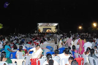 Night outdoor gathering of a large crowd seated on rows of white and red and blue plastic chairs, facing a lit open-air stage with a white canopy. On the stage, several robed figures in saffron and white stand near a central backdrop. In the foreground a woman in a white sari with gold border leans forward between the chairs, and nearby women wear pink, teal and green saris. Many attendees wear white. Lamps light the pathway at right.