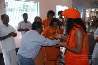 Indoor photograph in a pink-walled hall with blue-framed windows, showing a devotee in a white shirt and grey trousers bending forward to offer something to a saffron-clad sannyasi. The sannyasi on the right wears an orange turban, long dark swept-back hair, a sleeveless orange robe and a rudraksha mala, and holds out both hands to receive the offering. Beside them stands a bearded man in an orange robe with a gold-coloured necklace and glasses, and a shaved-headed man in orange behind. Several men in white dhotis and shirts stand around, some smiling and clapping, and a young man in an orange shawl with a camera watches nearby. The scene appears to be a ritual welcome or offering at a temple or ashram hall.