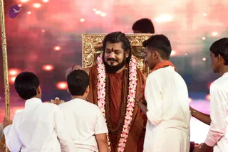 A seated man in rust-orange robes sits on an ornate gold throne on a stage with a pink-red glowing backdrop. He wears a long garland of white and pink flowers, a rudraksha mala, a red tilak on his forehead, and a gold necklace. Four young men in white kurtas and dhotis stand around him, two with hands near his shoulders and one holding a gold-tipped staff on the left. A gold ceremonial staff stands at the left edge. A probable Swamiji is seated in the centre, with long swept-back hair and orange robes, while others attend him in white ritual clothing.