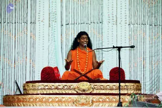 A long-haired man in saffron-orange robes and a multi-strand gold-beaded necklace sits cross-legged on an ornate gold-and-maroon throne in front of a white drape hung with beaded strings. He has open, animated hand gestures and is speaking into a microphone on a stand. A small red tambourine-like drum sits beside him, and a tiger-striped cushion rests on each side of the seat.