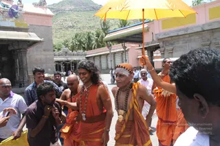 Outdoor photograph of a procession at a temple complex with pink and yellow walls, a mountain and palm trees behind. A bald man in orange robes with a white forehead band and a dark rudraksha mala, wearing a gold pendant, walks centre-right smiling beside a long-haired person in orange robes with a red-and-gold waist sash and layered beaded necklaces. An attendant holds a bright yellow umbrella over them. Other men in orange and white dhotis and striped shirts crowd around, and one man in a maroon shirt holds a yellow bag. The whole group moves along a paved courtyard.