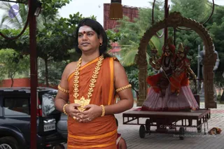 A standing man in an orange saffron dhoti and shawl with gold-yellow borders wears a long gold necklace with a large pendant, gold armlets and bangles, and a red bindi with tilak on his forehead; his long dark hair falls past his shoulders and his hands are folded at his waist. Behind him stands a parked dark SUV, and to the right a decorated ratha-style wooden arch frames two colourful deity murti figures, one in red and one in pink-white, on a wheeled wooden cart. Trees and palms fill the background. Swamiji (probable) is shown in appearance-based cues of long hair, saffron clothing and ornate gold jewellery, standing in an outdoor courtyard.