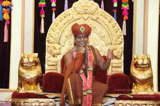 A seated person on an ornate gold throne with a carved golden back and two gold lion-head armrests draped in red tiger-striped cushions. The person wears an orange-red turban with a green and gold jewelled emblem, an orange-red robe, a long pink-and-white flower garland, rudraksha-style beads and gold bangles, smiling with one hand raised near the chin and the other gesturing. Colourful tassels hang at the top corners and a dark maroon drape forms the backdrop.