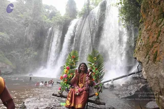 A seated figure in a reddish-orange silk dress with gold border sits on a wooden swing decorated with green palm fronds and pink, orange and yellow flowers, suspended before a tall waterfall in a lush jungle setting. A long multi-strand garland of flowers and a gold necklace hang around the neck, and the figure raises one hand in greeting while holding a small object. Water is visible in the foreground pool, with several people bathing in the shallows at left. A rocky orange cliff face rises on the right, and mist fills the air around the falls. The figure shows distinctive long, swept-back hair and ornate gold adornment, consistent with a probable appearance of a person seated in a ceremonial setting.
