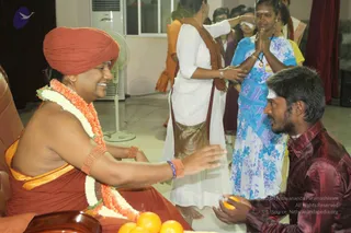A seated figure in a rust-red turban, orange-and-white flower garland and a saffron shawl smiles while extending a hand toward a kneeling young man in a maroon satin shirt who holds an orange fruit in cupped hands. Behind them, women in white and blue floral dress stand with palms pressed together, and another woman in white applies a white mark to a devotee's forehead. Oranges sit on a tray in front, and a pedestal fan and cream wall sit in the background. a person appears in the seated figure: a turban, saffron clothing and a flower garland, with a visible ritual bearing. The kneeling man offers fruit as a devotee, and the standing women are devotees and volunteers in a satsang-style gathering.