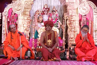 Three seated men in saffron and orange robes sit on low wooden chairs on a red-patterned rug before an ornate golden shrine. The central man wears a red turban, a rudraksha mala and a large ceremonial gold pendant necklace, and smiles with hands clasped in his lap; the figure on the left has a grey-haired shaved head with tripundra markings and speaks into a microphone on a stand, while the man on the right, with a long white beard and tripundra, joins his palms in greeting. Behind them stand deity murti with a trident, garlands of pink and white flowers, and kalasha-style lamp stands. The scene reads as a satsang or formal reception.