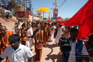 A large procession of sadhus moves along a stone ghat path under bright sunshine at the Ujjain Kumbh Mela. Many sadhus wear saffron and orange dhotis and shawls with long garlands of gold beads and pendants. A central sadhu in a rust-brown robe and orange turban walks at the front, and others in white dhotis follow behind. A large red flag and a yellow ceremonial umbrella are carried above the group, while a uniformed policeman walks beside them. A cameraman in a dark cap and blue vest films the procession on the right. Billboards and banners with portrait photos line the route, and a tiled staircase rises on the left.