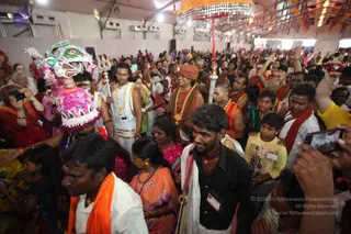 A photograph of a crowded indoor festival hall with a white tent ceiling and hanging decorations. In the foreground, a decorated ceremonial headdress with a colourful peacock-like figure and pink flowers is carried on a pole. A man in a white dhoti with gold necklaces holds a long silver staff with a trident-shaped top. Nearby, a woman in a saffron-orange sari and gold garland stands beside a man with a shaved head in orange wrap and gold chains. Many devotees in orange, red and white clothing surround them, several taking photos with phones and cameras. A large ornate umbrella-like canopy hangs above the crowd.