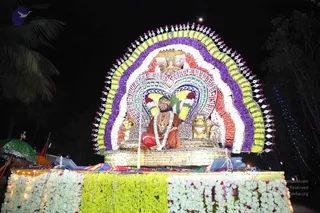 Night procession float decorated with garlands of white, yellow, purple and orange flowers arranged in a large arched backdrop with a radiating halo-like frame. A seated figure in saffron-orange and red robes, wearing a crown-like headpiece, a gold necklace and a flower garland, sits on a gilded throne flanked by two golden lion sculptures. Flower-covered panels and fairy lights decorate the float base, with a green decorated umbrella at the left edge and a palm tree silhouette against the dark sky.