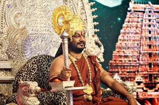 A man seated on an ornate silver throne with leopard-print cushions, wearing a gold crown with a tall gold ornament, a rust-orange draped garment, a gold chain necklace, a long gold rudraksha-style mala, and beaded gold bangles on his upper arms. He holds a tall silver staff with a trident-like top and a vibhuti or kumkum mark on his forehead. Behind him stands a large temple tower with red and orange sculpted figures under a dark starry backdrop. Swamiji (probable) is a seated figure with distinctive ceremonial adornments, crown and rudraksha-style mala, shown in person. Copyright text appears in the lower right corner.