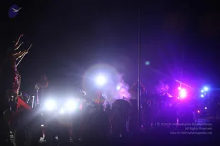 A night procession photographed in dark surroundings, lit by bright white and magenta spotlights and haze. Several people in dark clothing stand in a crowd along the right side, some holding umbrellas. On the left, a large ornate float or vehicle with decorative fringed structures and tall poles or staffs rises above the crowd, with red and orange flags and banners fluttering nearby. Smoke or incense haze drifts through the scene.