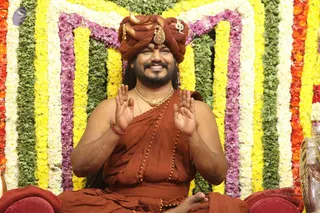 A smiling man seated cross-legged in front of a backdrop of vertical garlands of yellow, white, pink-purple and green flowers. He wears a rust-orange turban with a gold ornament and a gold crescent-shaped crest, an ornate gold necklace, rudraksha beads, and an orange-brown ochre cloth draped over bare shoulders. His palms are raised in greeting or blessing. a person: long hair and an ornate turban and gold ornaments, consistent with the discourse setting.