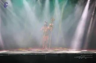 A performer stands alone on a stage in haze lit by beams of green and white light. He wears a Shiva-style costume with a crescent-like headpiece, a dark ornamented headband, reddish-orange body paint, and a waist cloth with gold details. He holds a tall golden trident in one hand and raises the other hand in a blessing gesture. This is a stage performance depicting a Shiva form, shown during a stage presentation at the event.