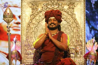 A man seated on an ornate throne with a gold-framed embossed backdrop, hands pressed together in anjali greeting and smiling. He wears a deep red turban, a red-orange cloth wrap over his shoulders, a rudraksha mala across his chest, and a gold necklace. Behind him is a leopard-print cloth on the seat. Beside him stand a gold staff topped with a red-jewelled finial and two ornate golden peacock figures. A large portrait of a man with a red tilak is visible on the left. Swamiji (probable): the seated man shows long swept-back hair beneath the turban, ornate gold necklace, rudraksha mala and orange-red robes, consistent with the page's reference to Swamiji at this event.