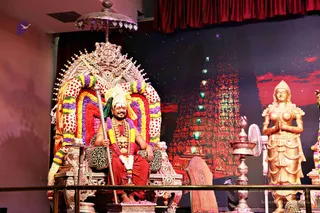 A seated man on an ornate silver throne with a floral-garlanded arch of red, yellow, purple and white blossoms. He wears a red dhoti, a leopard-print cloth on the seat, a gold ceremonial necklace, a rudraksha-style mala of flowers and a turban-like headdress, and holds a silver ceremonial staff. Beside him stands a golden statue of a female figure with hands folded in namaskar, and a silver lamp stand and fan sit at the right. The backdrop shows a red illuminated temple gopuram with a starry sky. A tall red curtain frames the stage. Whether this is the figure named on the page is uncertain from appearance alone, so the identity is marked unsure.