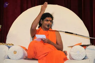 A photograph of a seated man in long flowing saffron-orange robes on a large white rounded throne against deep red velvet drapery. He has long dark hair swept back, a small red bindi on his forehead, and his right arm is raised high in a gesture while he holds a sheaf of white papers in his left hand. A microphone on a flexible stand is in front of him, and white bolster cushions flank the throne. A gold ceremonial item is visible at the left edge. Appearance-based candidate, probable Swamiji, seated on the throne delivering a discourse, identified by long swept-back hair, saffron robes and a red bindi.