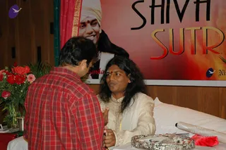 Indoor event photo at a Shiva Sutras session. A man in a red and black plaid shirt, seen from behind on the left, leans toward a long-haired man seated on a white-draped bench with a red bindi, wearing a cream embroidered kurta and white shawl; the seated man gestures with raised hands in conversation. A silver tray holding flowers sits in front. On the left, a vase of red roses and pink gerberas stands on a table. Behind them a large red-and-gold banner reads SHIVA SUTRA and shows a smiling man in a white turban and dark jacket. Two people are visible in the scene.