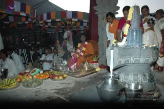 Photo of a Shiva lingam abhishekam at an open-sided pavilion with striped fabric canopy. A large grey stone lingam stands on a carved yoni-shaped base, draped with white and yellow flower garlands; a woman in a red blouse and white sari pours from a small pot while another woman in a white sari holds a metal pot. A man in white and orange, with a brass trident beside him, stands at the base. Seated in front, a person in orange robes wears a red-and-white flower garland, with brass lamps, a metal vessel, and trays of bananas, oranges, apples and custard apples in front. Steel vessels catch the flowing water at the bottom, and the crowd sits behind.