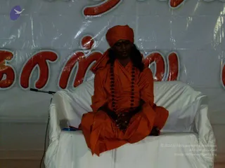 A photograph of a single man seated cross-legged on a white-draped sofa at a stage or indoor event. He wears an orange-saffron robe and an orange turban, with a long rudraksha mala hanging across his chest and his hands resting together in his lap. Behind him is a white vinyl banner with large red-orange Tamil lettering. A microphone stand is visible to the left. Probable Swamiji, seated in saffron robes with a rudraksha mala and turban, giving a discourse at the Attur event.