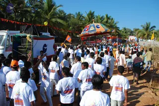 A large crowd walks down a dirt road lined with coconut palms during a procession. Most participants wear white T-shirts printed with colourful yoga-figure designs, and some wear white dhotis or kurtas. A large umbrella with blue, red and yellow panels and a golden finial is held over the procession. A decorated white vehicle with a green canopy and loudspeakers, strung with red and yellow bunting, travels alongside. Orange and yellow flags flutter over the road, and a balloon floats near the vehicle.