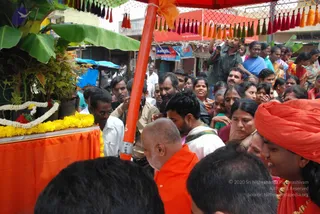 Outdoor street procession in a crowded Indian town. A bald, bearded man in an orange shawl stands at centre-front, surrounded by devotees; a woman at right wears a bright orange turban and orange garment with a gold necklace. Behind a red umbrella hung with orange and red tassels, a crowd of men and women in saffron, white and colourful saris presses forward. At left a decorated orange-draped platform holds potted plants with yellow marigold garlands and white flower strings. A man in white shirt and dhoti stands at left, and a cameraman with a camera is visible behind the crowd. Shop signs and an Aircel advertisement line the street.