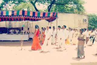Outdoor street scene in a dusty courtyard beside a whitewashed wall and trees. Beside and behind them, a bald man in white shirt and white dhoti walks barefoot, and several men in white shirts and white dhotis follow, some with yellow cloth. Women in white and cream saris walk among them. A striped blue, green and pink canopy shelters a stall on the left where people in white sit behind a table with offerings. On the right, an older man in a khaki shirt and checked lungi stands watching. Visible people number about 20, including those partially shown.