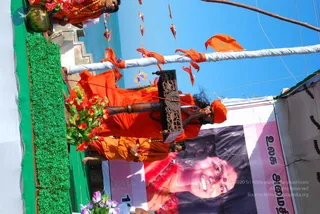 Photograph of a decorated float at an outdoor procession under a clear blue sky. A figure in orange saffron robes and an orange head wrap stands on the float, holding a long wooden staff with a carved ornate ornament, blowing a conch or horn-like instrument to their mouth. Another person in orange robes stands behind, and a third figure in orange is visible at the top edge. The float is lined with green artificial foliage and red flowers, and orange triangular pennants hang on strings above. A large banner beneath shows a smiling woman in red with a red bindi, with a 2009 date printed on the banner. Tamil text appears on the banner.