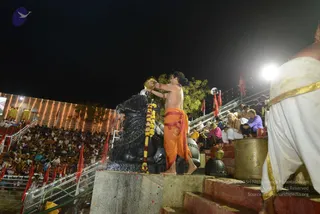 Night photograph of an outdoor abhishekam at an illuminated ghat-style stepped amphitheatre. A bare-chested man in an orange dhoti and orange cloth sash pours liquid from a golden vessel over a large black stone lingam, which is draped with a yellow and orange marigold garland. Behind him, rows of seated devotees fill the steps and stands under strings of lights, with red and saffron flags along the railings. On the right foreground, a man in a white dhoti with gold border stands near a large brass pot. Bright floodlights and red-and-white trim complete the scene.