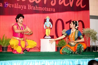 Stage performance at a festival event with a red backdrop reading "Brahmotsava" and "2010", with partial text "Devalaya" and "Nithyanandeshwara". Two dancers kneel on a green stage: a man on the left wears a pink sleeveless top, yellow dhoti with a gold belt, rudraksha mala and a mustache, gesturing with his hands; a woman on the right wears an orange sari with green border and blue blouse, flowers in her hair, pearl necklace and bangles, extending one arm toward him. Between them stands a blue-skinned deity murti of Krishna with a crown, yellow dhoti and garland, on a white pedestal. Potted plants frame the stage edges.