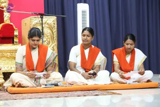 Three women sit cross-legged on an orange cushion in front of a golden ornate throne and a red-cushioned seat. Each wears a white sari with coloured borders and an orange shawl draped over the shoulders. The woman on the left and the one on the right read printed sheets of paper, while the woman in the middle holds a black microphone and looks down. A standing microphone stands between them and the throne, with a blue curtain and a white tower fan behind.