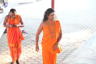 Two men walk across a hexagonal paved courtyard under a covered walkway near a white vehicle. The foreground man has long swept-back dark hair, wears a ceremonial gold bead necklace and gold armlets and bangles, and is draped in saffron-orange cloth. Beside him, a second man in orange robes with a yellow border looks down at a mobile phone he holds in both hands, with a rudraksha mala visible on his wrist. Sandals sit on the pavement, and a red pillar stands at the edge of the frame.