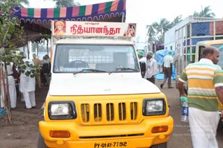 A yellow utility van with a white cab and open cargo bed stands at a relief camp in front of a striped purple, orange and green canopy. A banner on the cab reads paramamurumar in Tamil script beside a portrait of a saffron-robed figure and the word Nithyananda in Tamil. Several people in white clothing and green medical caps move around the site, including a man in a striped green and yellow shirt at right holding a water bottle. Palm trees and a white building stand behind, with a blue tarp on the right.