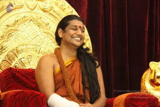 A smiling man with long black hair swept back sits on a red tiger-striped cushioned throne in front of an ornate gold carved backrest and deep red velvet curtains. He wears an orange saffron wrap with a yellow edge, a heavy braided gold-toned necklace, and a small red bindi on his forehead. His left forearm is wrapped in white bandage. Gilded lion-head armrest detail is visible at right. Swamiji (probable): long swept-back hair, saffron clothing, ceremonial gold necklace and throne seating are the qualifying cues, and the figure is the seated main subject. Single physical person in a formal satsang-style setting.