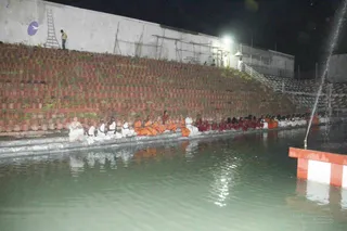 Night photograph of a rectangular pool with a stepped terraced bank of terracotta pots behind it. A long row of seated people in orange and maroon robes and white dhotis sits along the pool's edge, facing the water. A spray fountain rises from the water at right beside a red-and-white striped post. Floodlights illuminate the scene and a few standing figures in white stand along a wall at top. A purple logo appears on the wall at upper left.