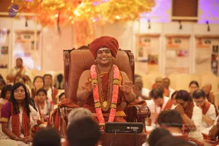 A seated figure in an ornate brown leather armchair sits centrally at a satsang-style gathering, wearing a deep red turban, a saffron-orange robe, a long gold-toned necklace with a pendant, and a thick pink and white flower garland. Both hands are raised in front of the chest with palms open, and a small headset microphone is visible near the face. Around the chair, a large seated audience in white and saffron-orange shawls sits on the floor, several with hands folded or eyes closed. A black Yamaha audio unit sits on a stand in front of the chair. Behind them, a warm orange-toned hall displays panels and framed images, with an illuminated tree-like backdrop.