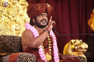 A man seated on a leopard-print cushioned chair before a golden ornate backdrop and red drapes, speaking into a microphone while raising his right index finger. He wears a rust-red turban with a small gold ornament, a red-brown saffron-toned cloth draped over his body, a long gold ceremonial necklace with beads and a pendant, and a pink and white flower garland. A golden lion-head sculpture sits beside him on the right. Swamiji (probable): long hair visible at the sides, gold necklaces and a saffron-toned cloth, appearing as the speaker in a discourse setting.